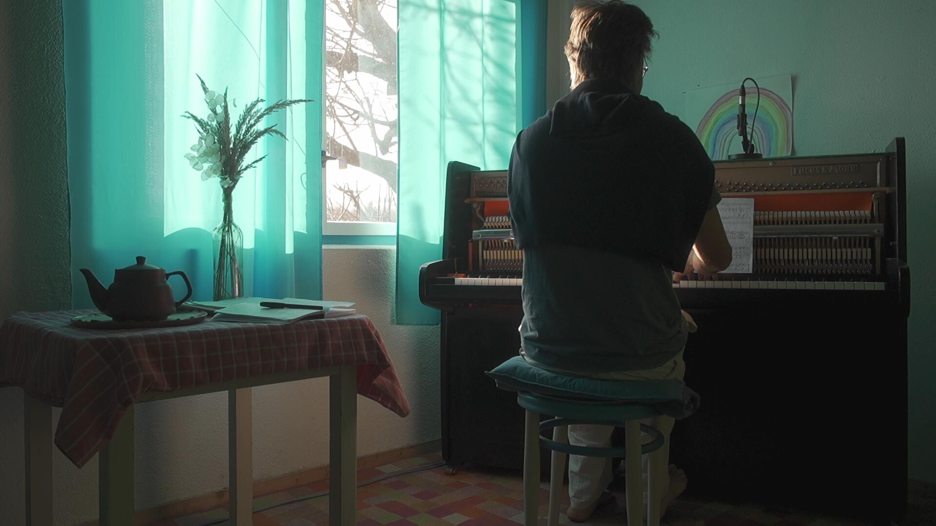 Musician seated at a piano bathed in soft natural light, surrounded by artistic elements.